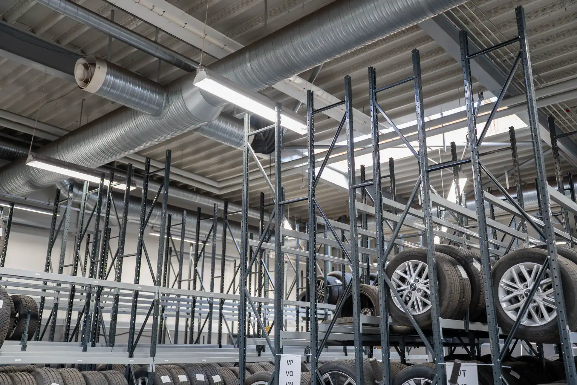 Billedet viser et lager med opbevaring af bildæk på metalreoler i en industriel setting.