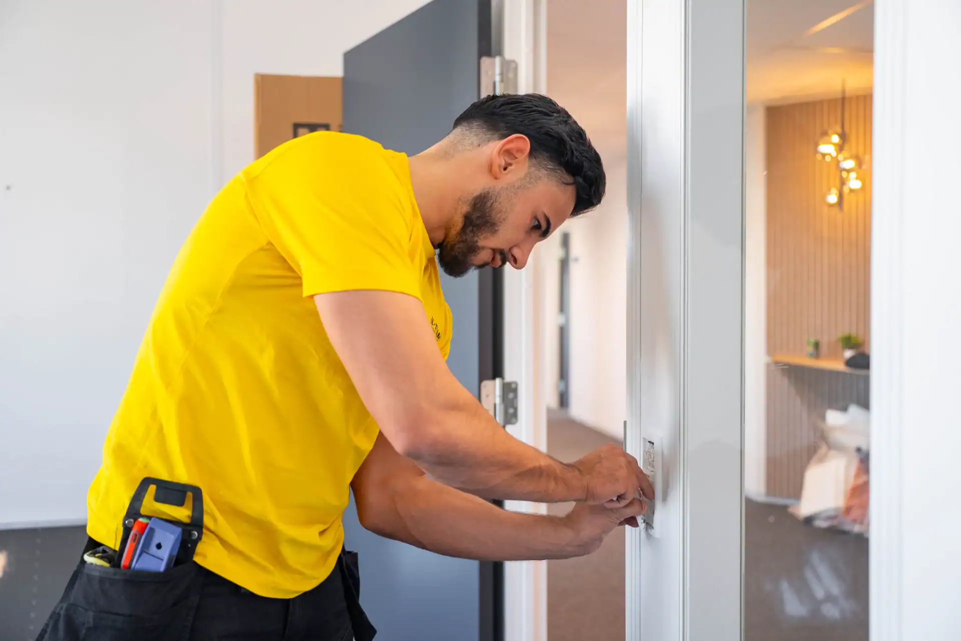 Billedet viser en mand i gul T-shirt, der arbejder med at montere eller reparere en dør.