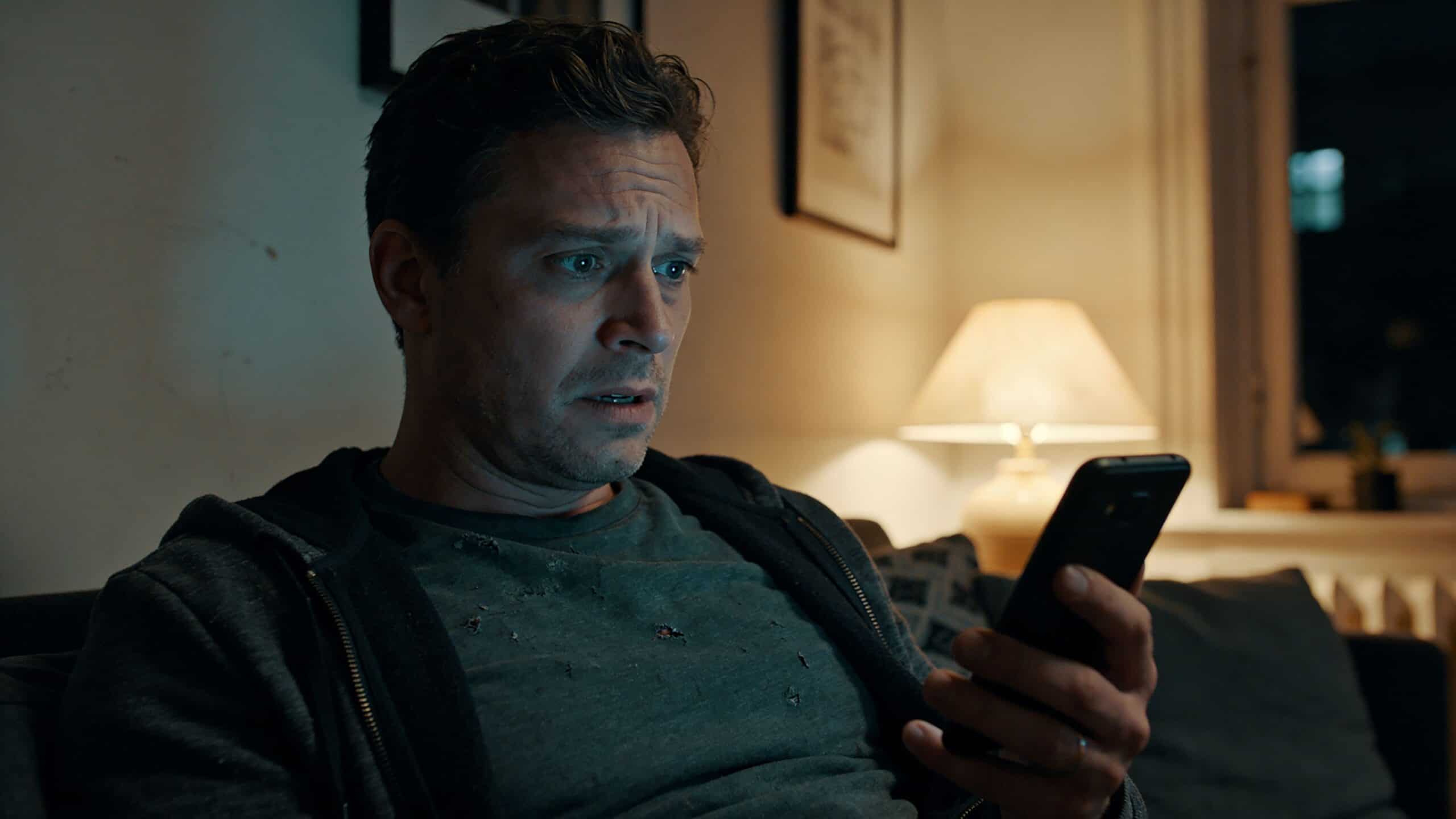 Mand tjekker sin smartphone, mens han sidder i stue med elektrisk udstyr i baggrunden.