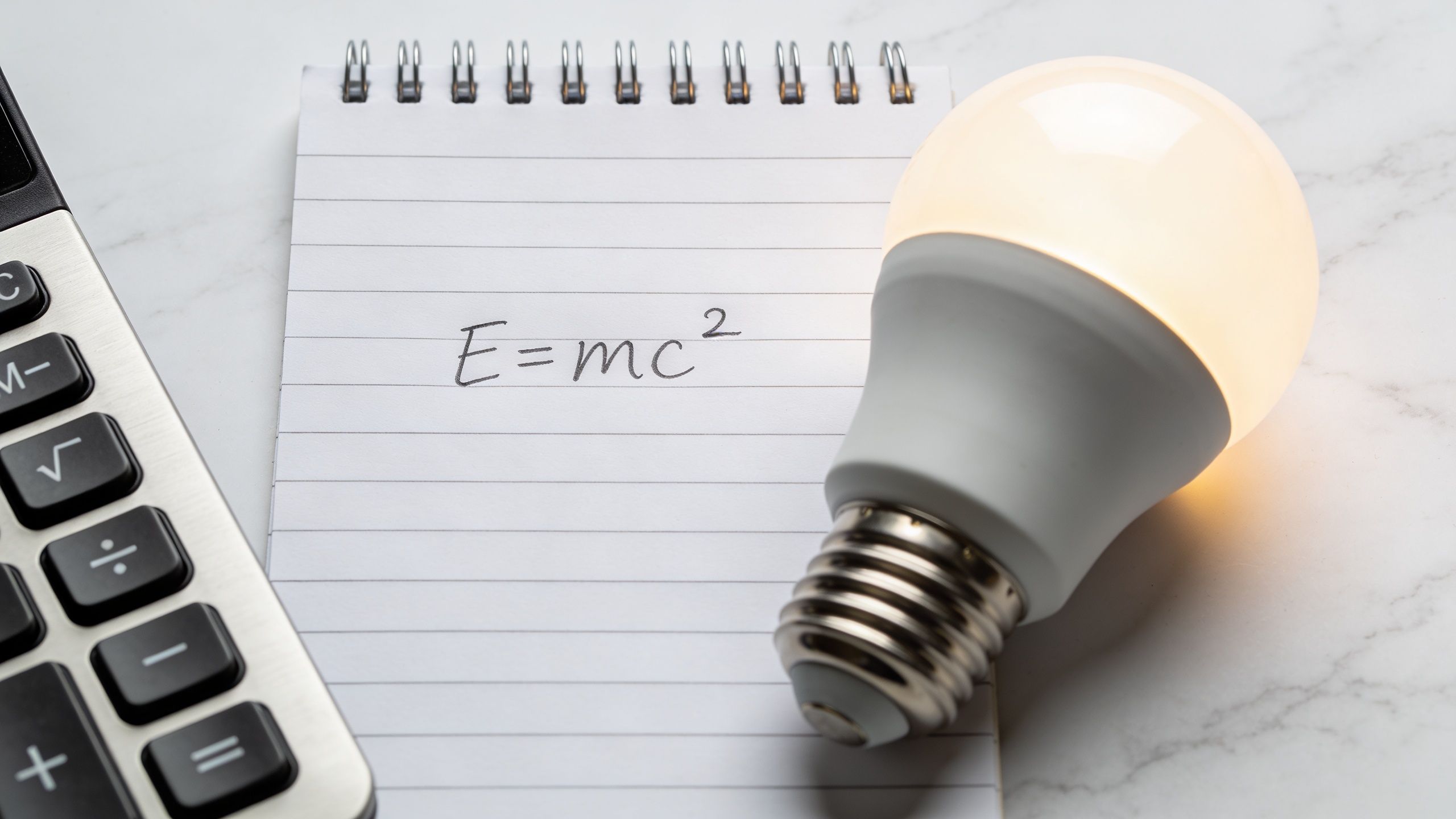 En lommeregner, en notesblok med formlen E=mc2 og en lysende elpære på et bord.
