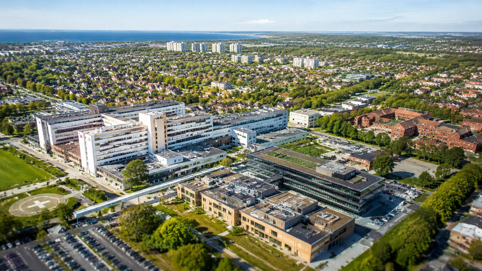 Luftfoto af et moderne hospitalskompleks med tilhørende bygninger og parkeringsområder i et grønt bylandskab ved kysten.